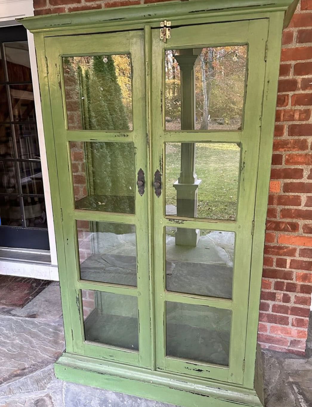 A vintage green wooden cabinet with glass doors stands on a stone porch. It has two handles and is against a brick wall. The cabinet is empty, and a reflection of the outdoors with trees and grass is visible in the glass.